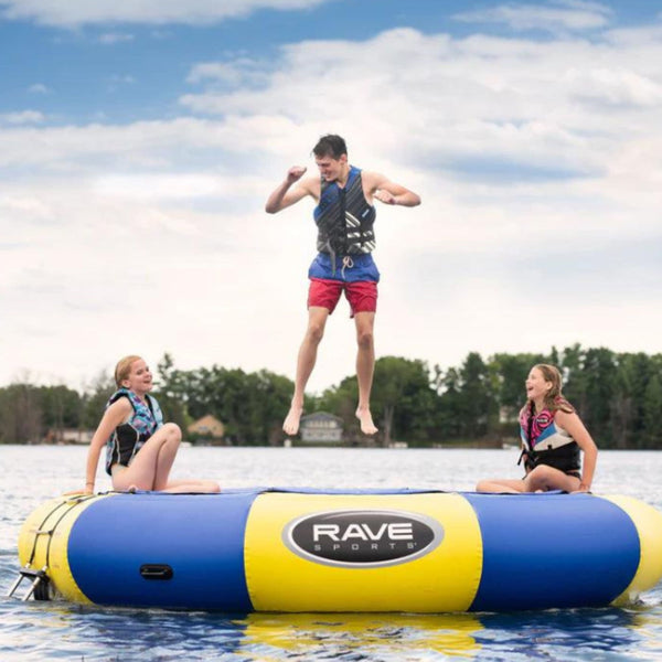 Blue and yellow Rave Sports inflatable water trampoline floating on a lake, with one person mid-jump and two others sitting on the edge.