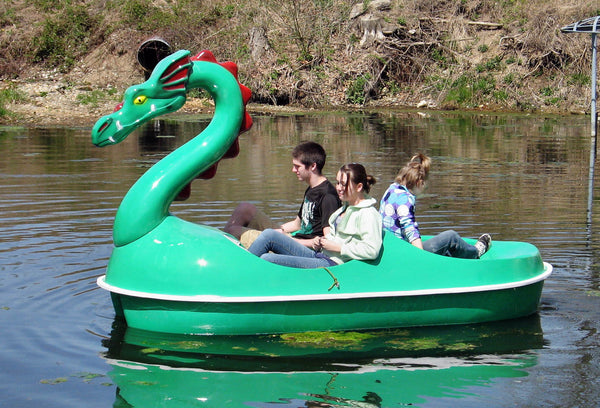 Green dragon-shaped pedal boat with a red-crested head, carrying three riders on a calm river