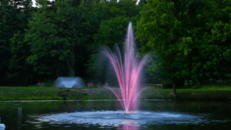 Pond fountain with pink LED lights creating a tall center plume and arcing sprays on a tree-lined lake at dusk.