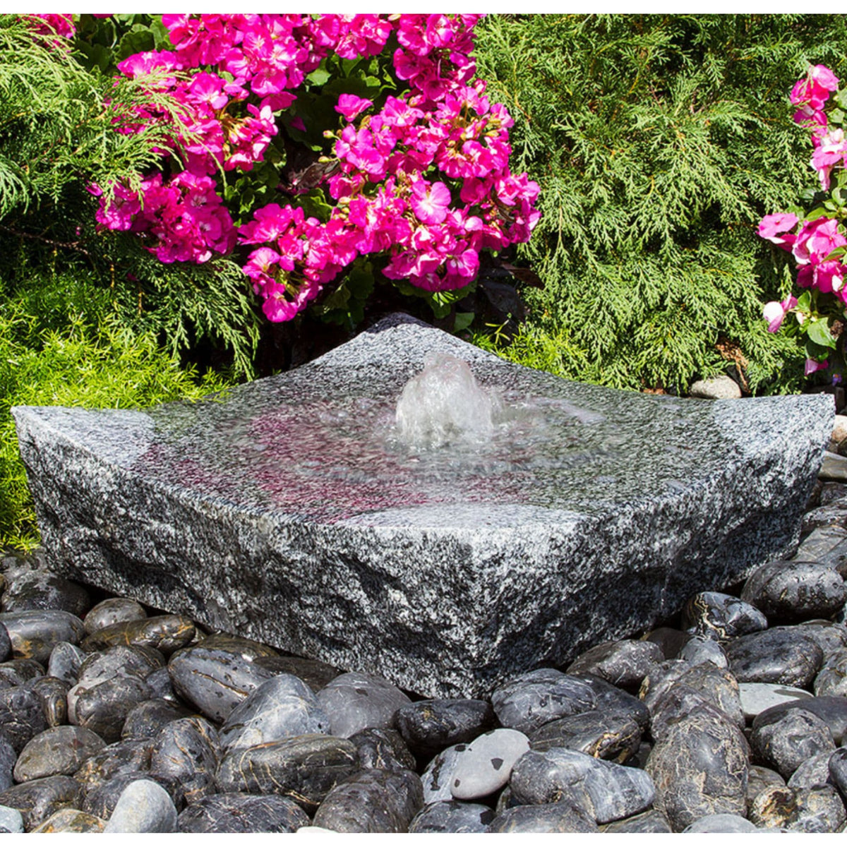 Blue Thumb bowled zen granite stone fountain holding a still pool of water among pink flowers and green foliage