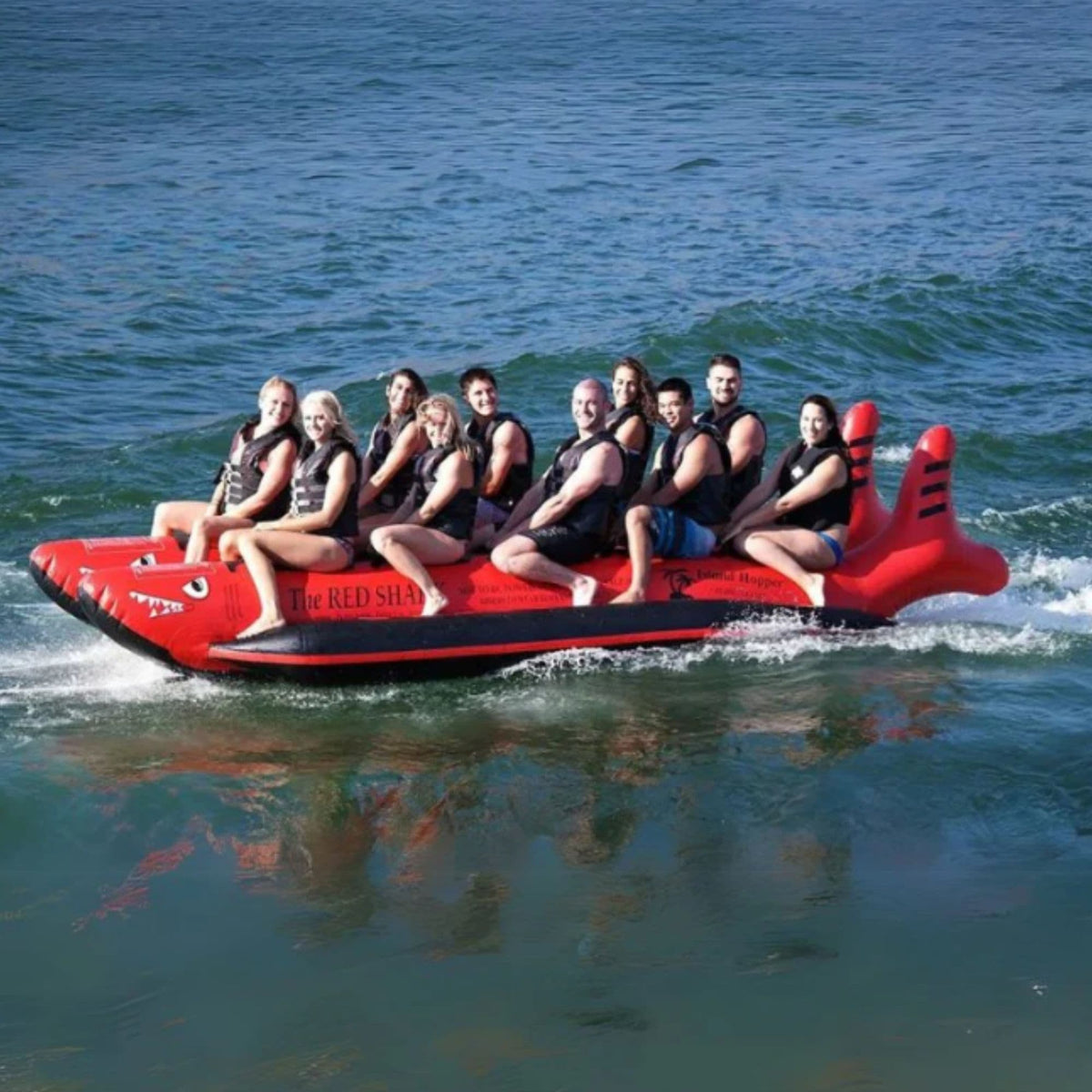 Ten riders in life jackets straddling a red shark-shaped banana boat as it is towed across choppy water