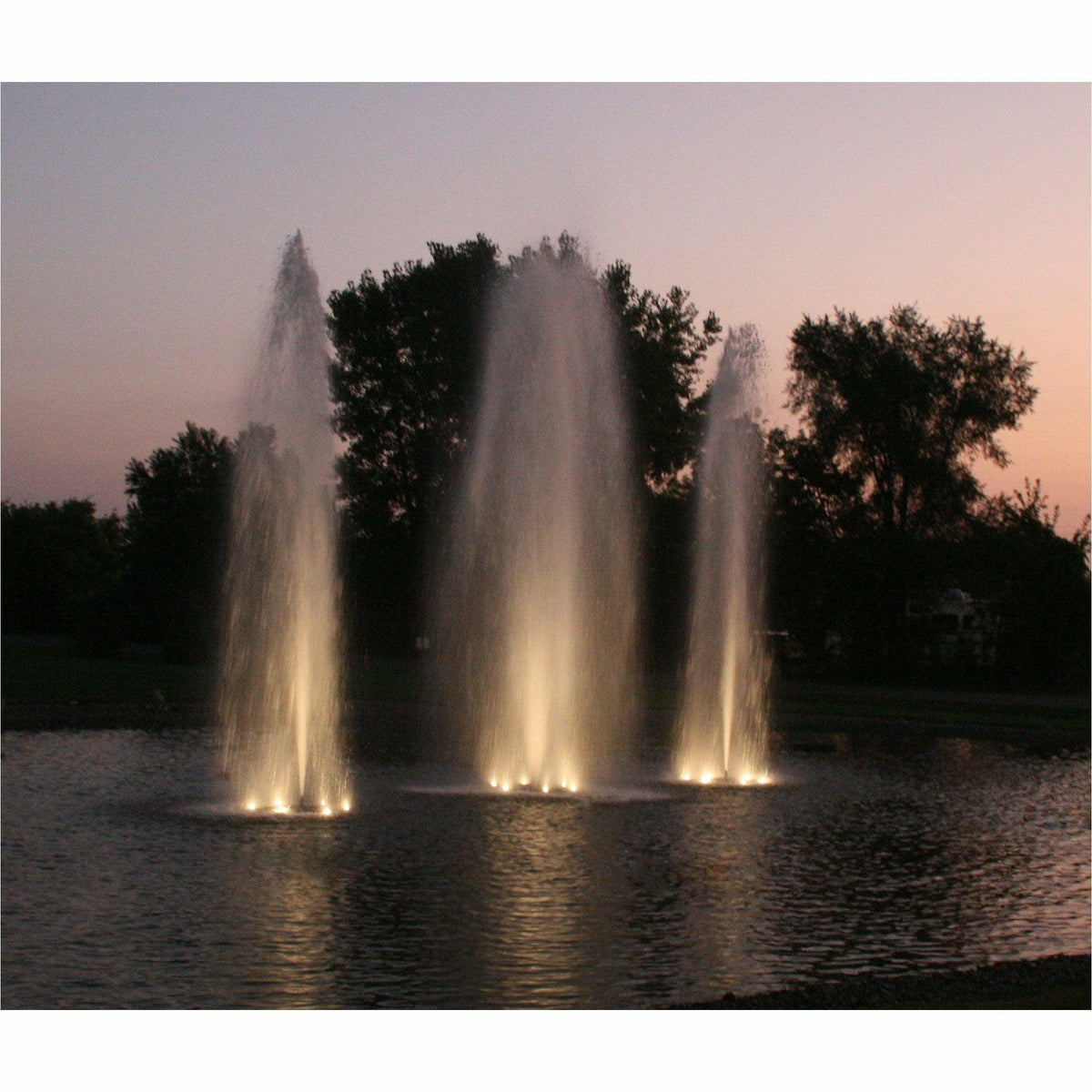 Three fountain plumes lit from below at dusk on a dark pond with a treeline behind