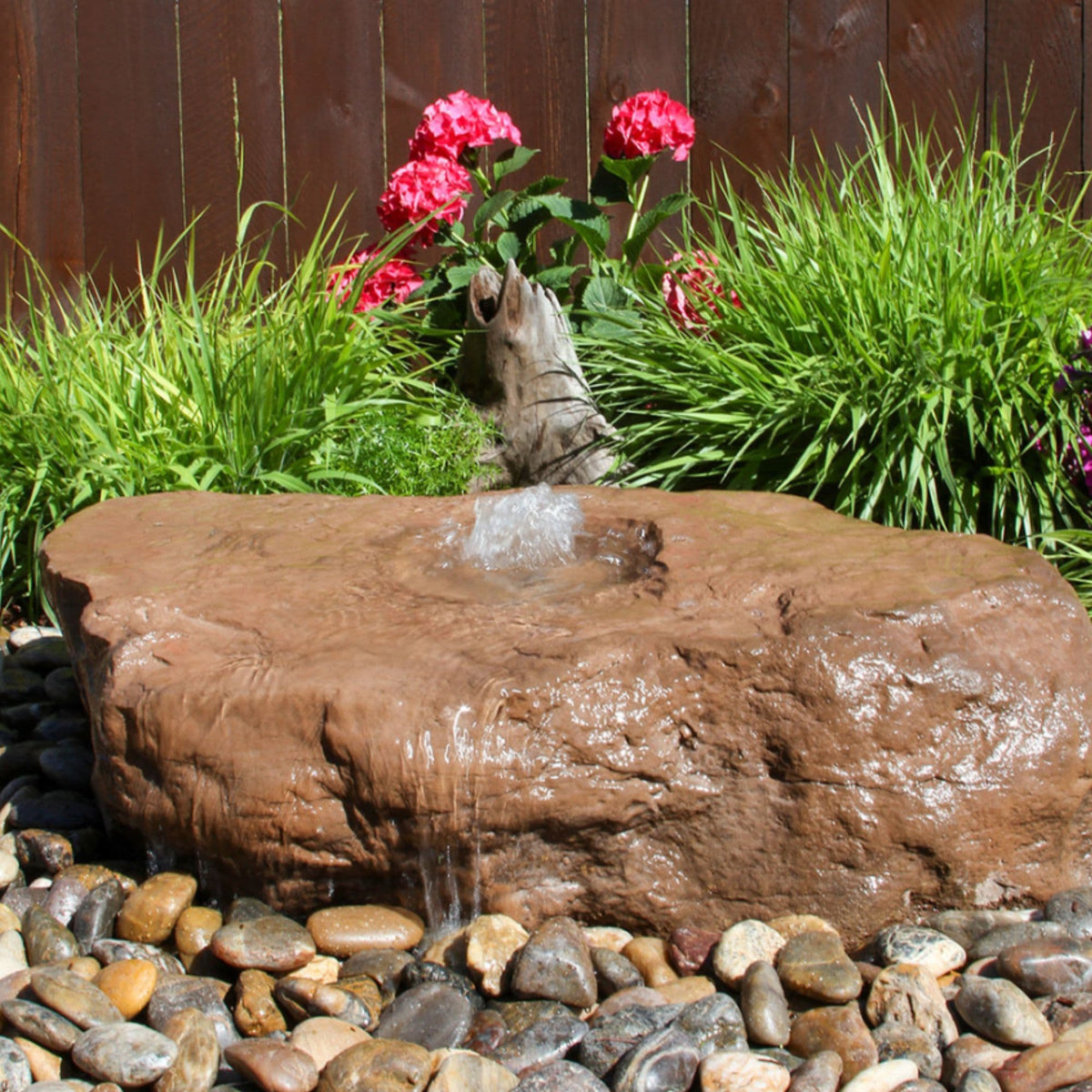 Blue Thumb large bird bath boulder stone fountain holding shallow water among ornamental grasses and flowers by a wooden fence