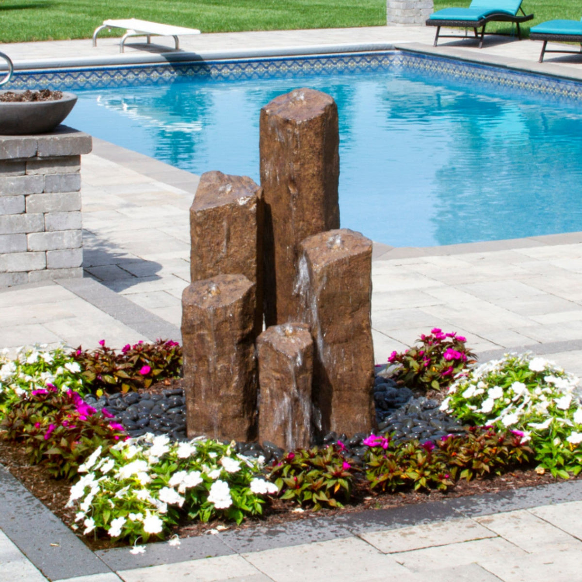 Blue Thumb Pent 5 column faux basalt fountain standing in a flower bed with a swimming pool behind it
