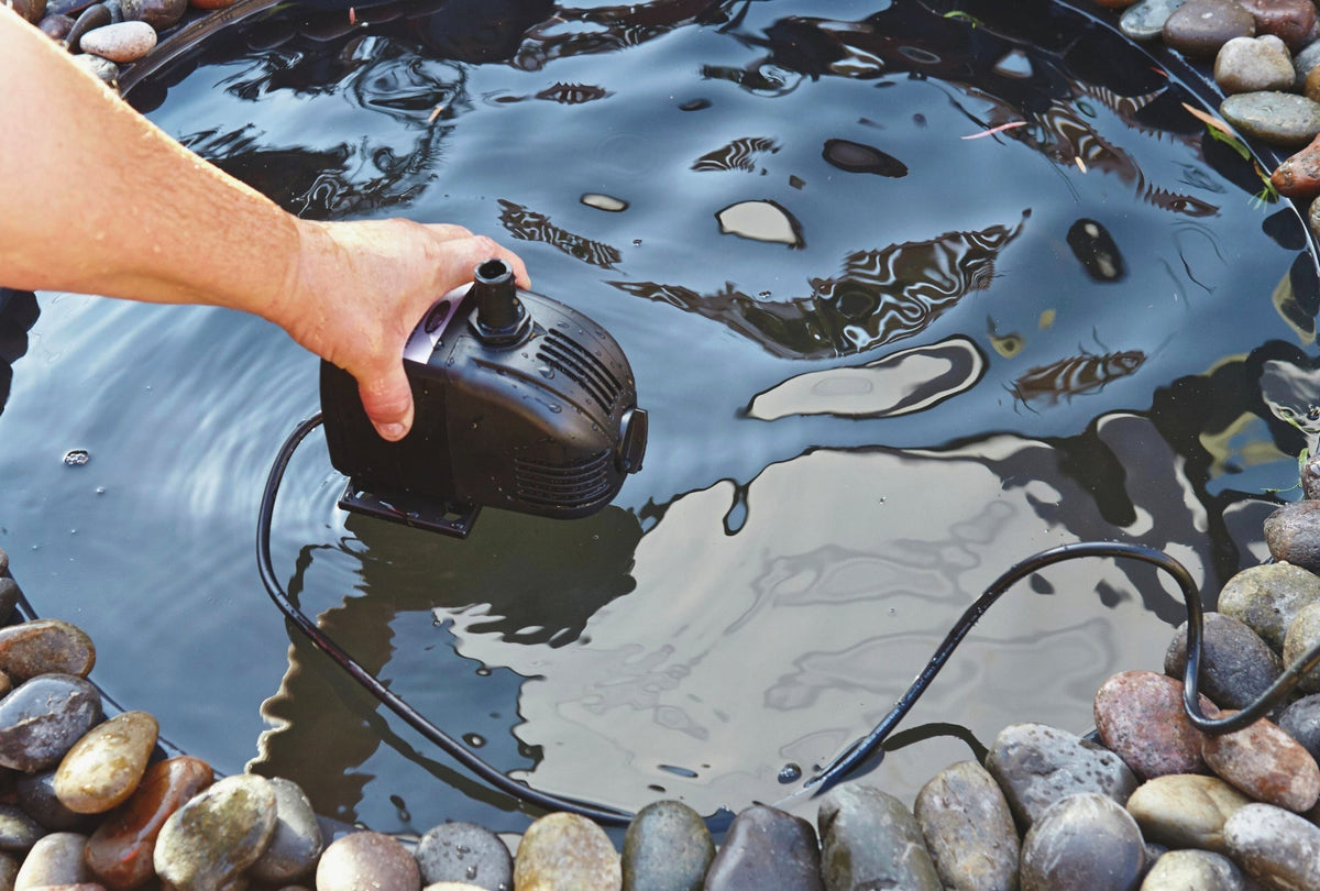 A hand lowering a small black submersible pond pump into a rock-edged garden pond.