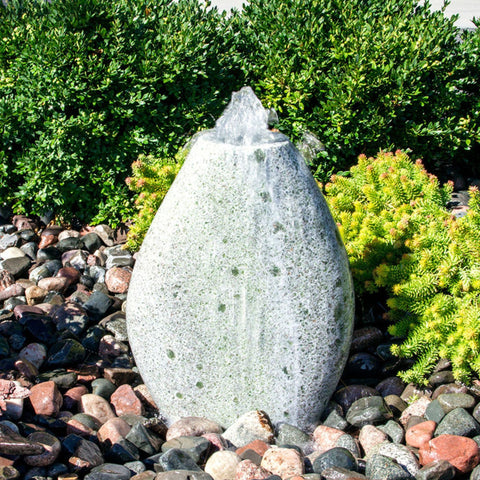 Almond-Shaped Green Marble Stone Fountain With  Small Trees Or Plants In The Background And Surrounded By Stones