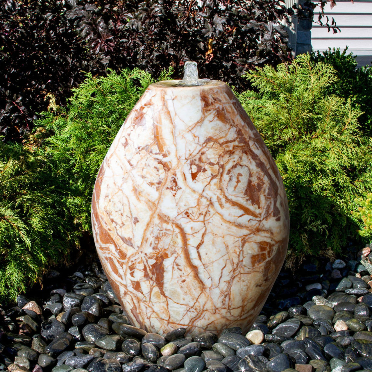 Almond-Shaped Red Marble Stone Fountain With Plants In The Background And Surrounded By Stones