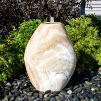 Thumbnail for Almond-Shaped Yellow Onyx Stone Fountain With Plants In The Background And Surrounded By Stones
