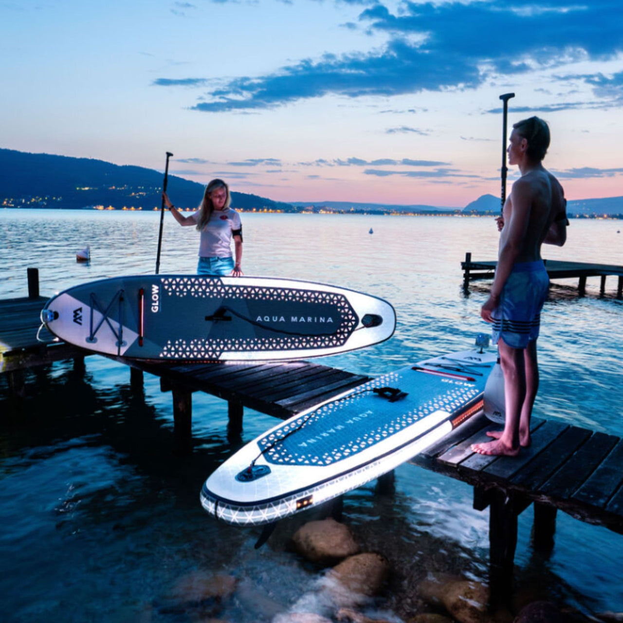 Aqua Marina Glow Paddle Board With A Man And Woman Standing And Getting Ready