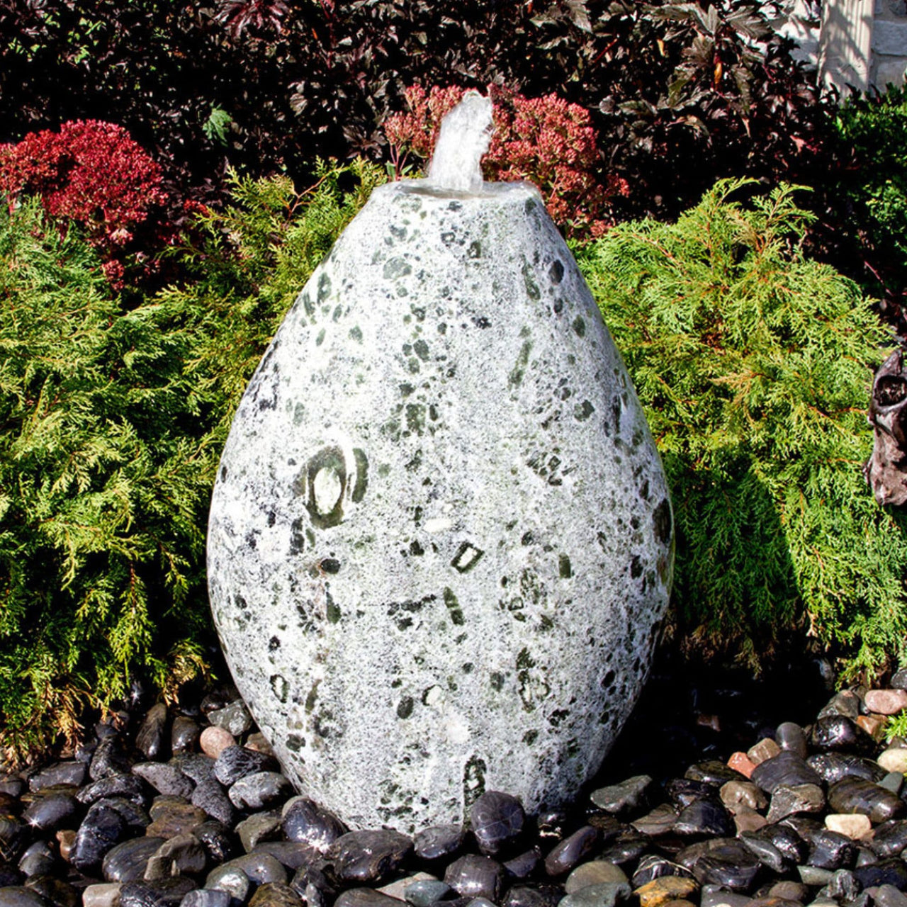 Blue Thumb Almond-Shaped Green Marble Stone Fountain With  Small Trees In The Background And Surrounded By Stones