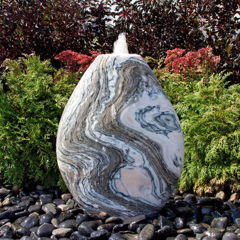 Blue Thumb Almond-Shaped Pink Marble Stone Fountain With  Small Trees In The Background And Surrounded By Stones
