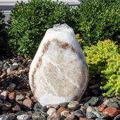Blue Thumb Almond-Shaped Red Onyx Stone Fountain With  Small Trees Or Plants In The Background And Surrounded By Stones