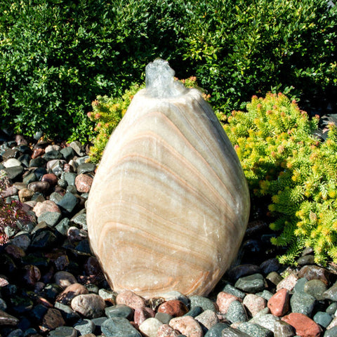 Blue Thumb Almond-Shaped Yellow Onyx Stone Fountain With  Small Trees Or Plants In The Background And Surrounded By Stones