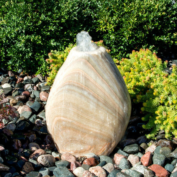 Blue Thumb Almond-Shaped Yellow Onyx Stone Fountain With  Small Trees Or Plants In The Background And Surrounded By Stones