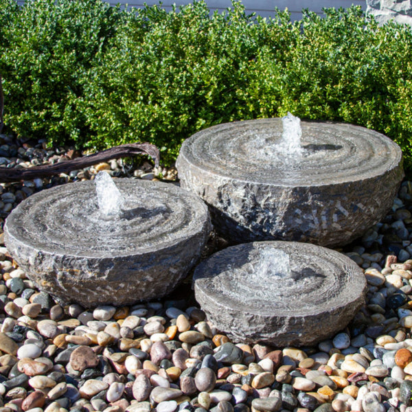 Blue Thumb Natural Stone Bowl with Swirl Fountains - Set of 3 Surrounded By Pebbles With Green Plants Behind