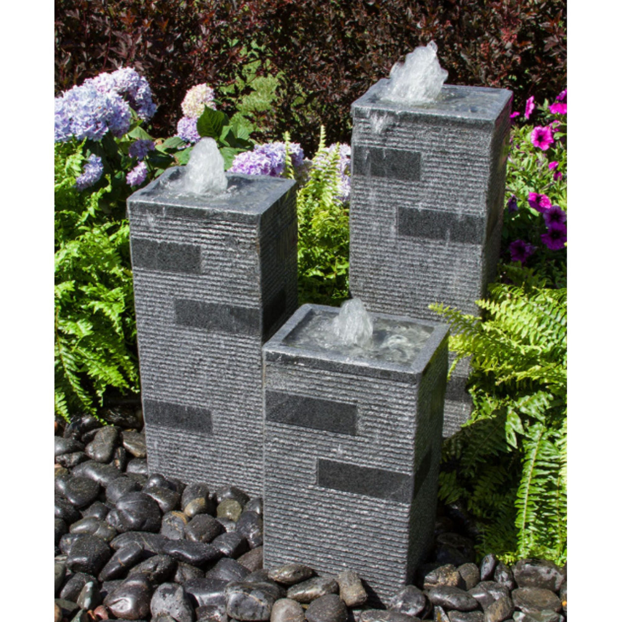 Blue Thumb Polished Block Stone Towers Outdoor Fountain Surrounded By Ferns And Flowers With Red Trees Behind