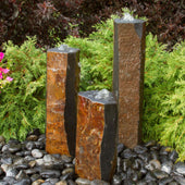 Blue Thumb Semi-Polished Basalt Rock Column Fountain Surrounded By Shrubs And Burgundy Leaves Plants
