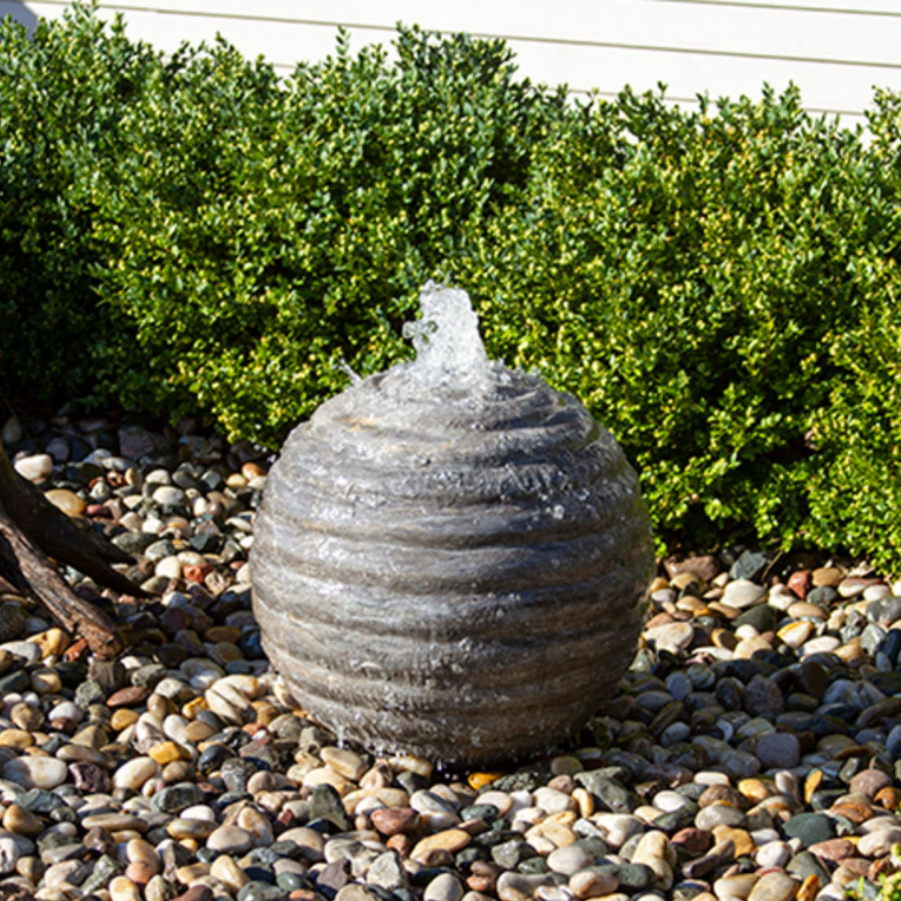Blue Thumb Smooth Ribbed Limestone Sphere Stone Fountain Surrounded By Green Plants With White Wall