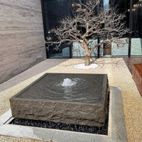 Thumbnail for Blue Thumb Yoshida Square Stone Fountain On Black Pebbles With A Tree In The Background