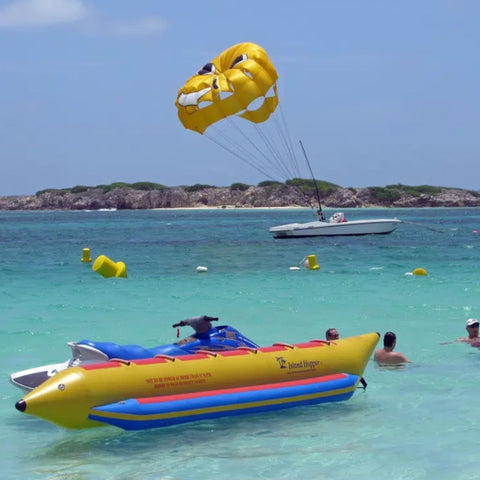 Elite Class 6 Person Banana Boat Resting In Clear Waters With Parasailing Over Three Men And Parasailing Over Nearby