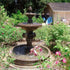 Thumbnail for Giannini Garden Quattro Lion Two Tier Pond Fountain Surrounded With Plants And A Small House In The Background