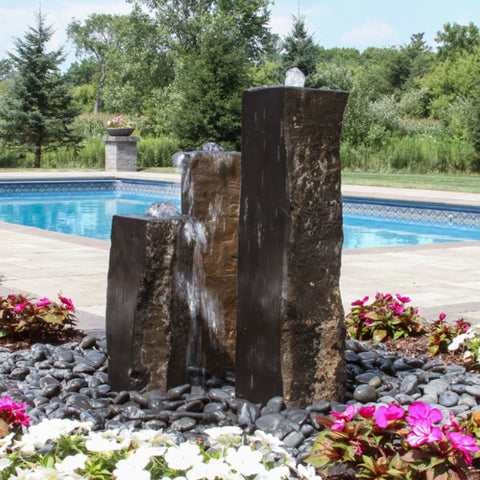 One Side Polished Basalt Stone Fountain Surrounded By Flowers With Pool And Trees Backdrop