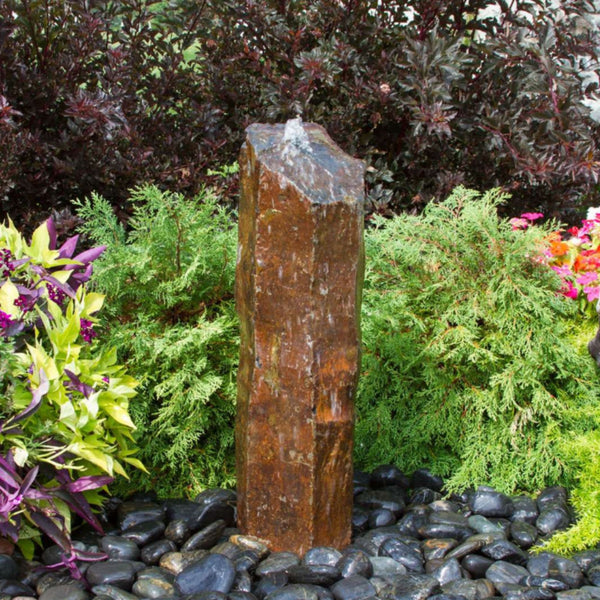 Outdoor Basalt Column Fountain Surrounded By Shrubs, Colorful Plants And Flowers