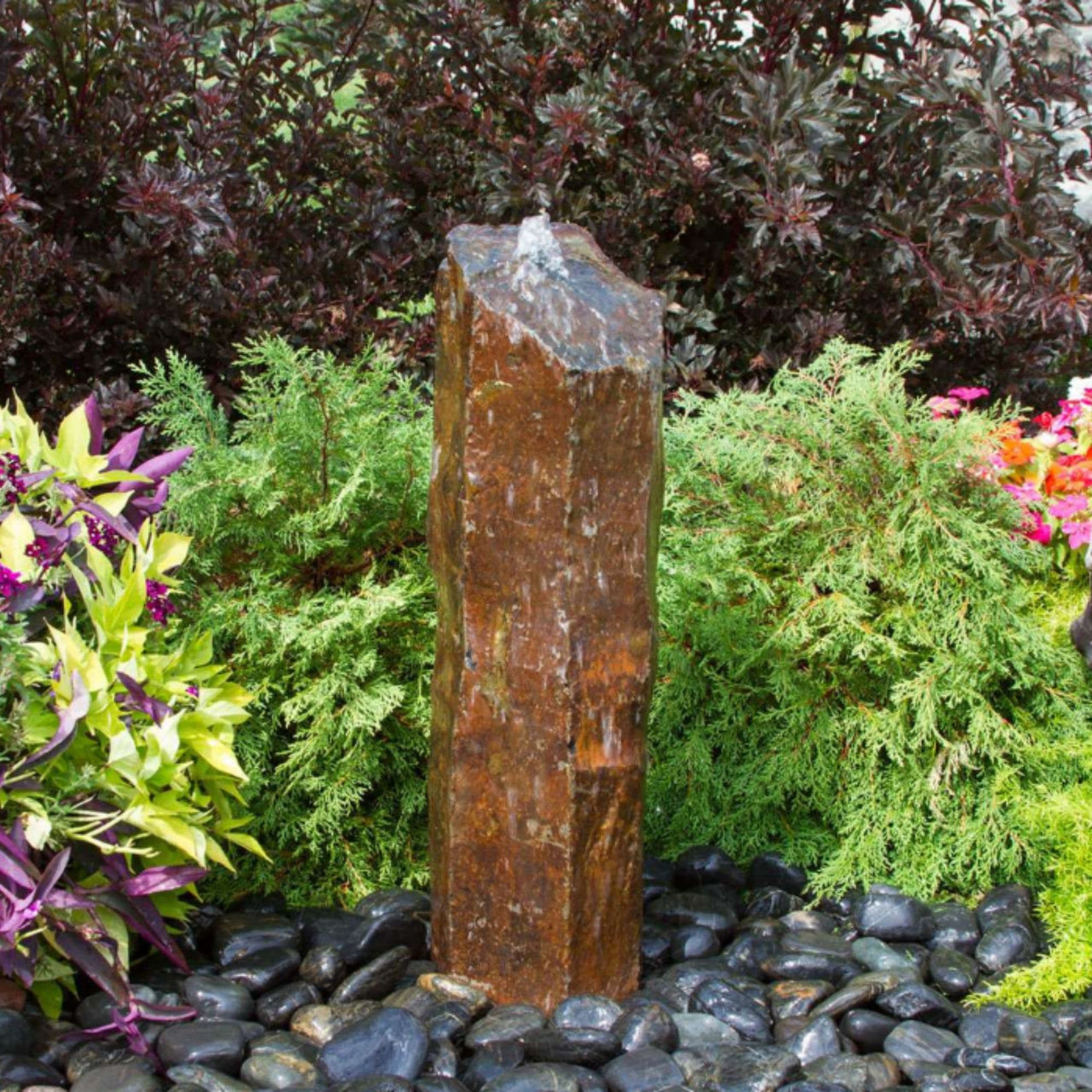 Outdoor Basalt Column Fountain Surrounded By Shrubs, Colorful Plants And Flowers