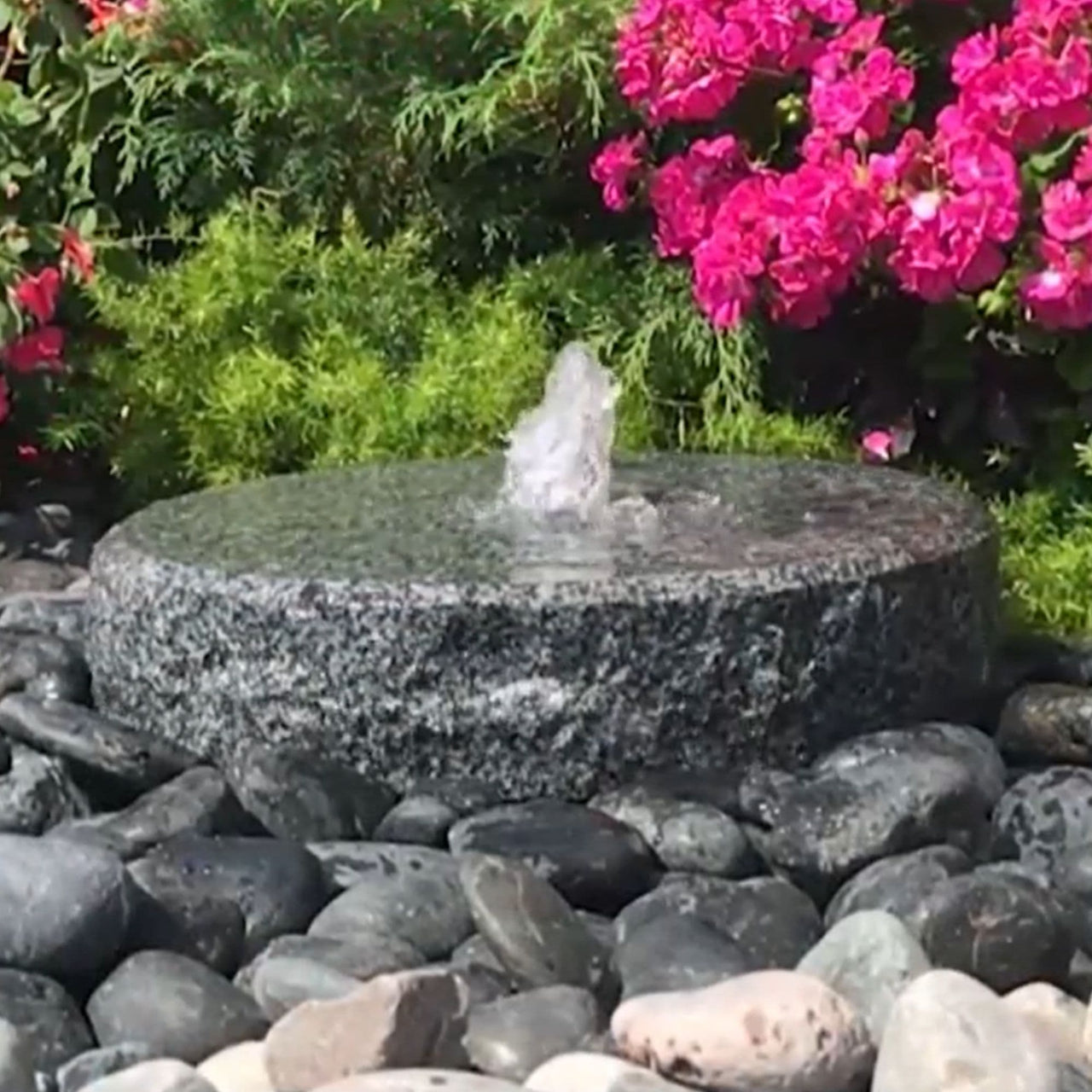 Outdoor Round Granite Fountain Surrounded By Pebbles With Shrub Plant And Flowers Behind