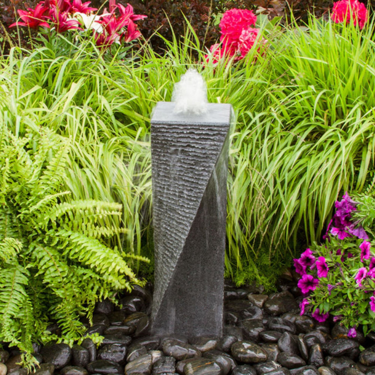 Polished Twist Granite Stone Fountain Surrounded By Ferns, Plants And Vibrant Flowers