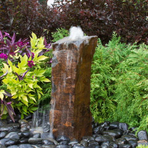 Rustic Basalt Rock Column Fountain With Shrubs And Colorful Plants Backdrop