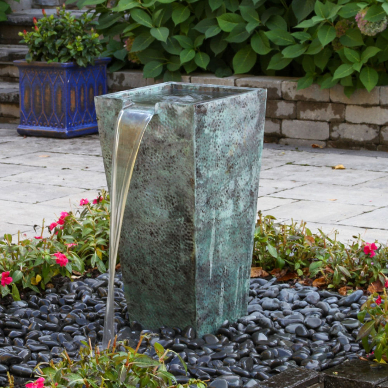 Single Tapered Square Urn Stone Fountain Surrounded By Ornamental Plants With Stone Backdrop