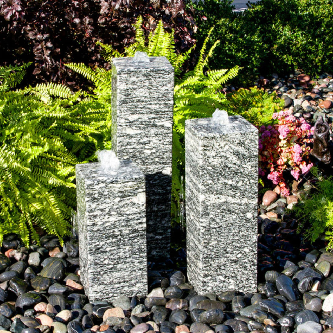 Smooth Speckled Granite Triple Stone Fountain Surrounded By Colorful Plants And Flowers Background