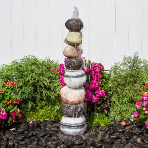 Stacked Pebble Garden Fountain Surrounded By Colorful Flowers And Shrubs Background With White Wall