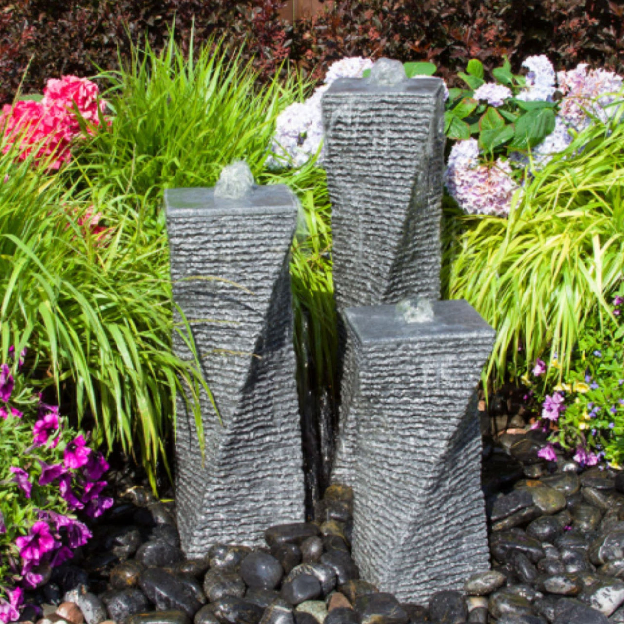 Triple Chiseled Twist Granite Stone Fountain With Colorful Plants And Flowers Background