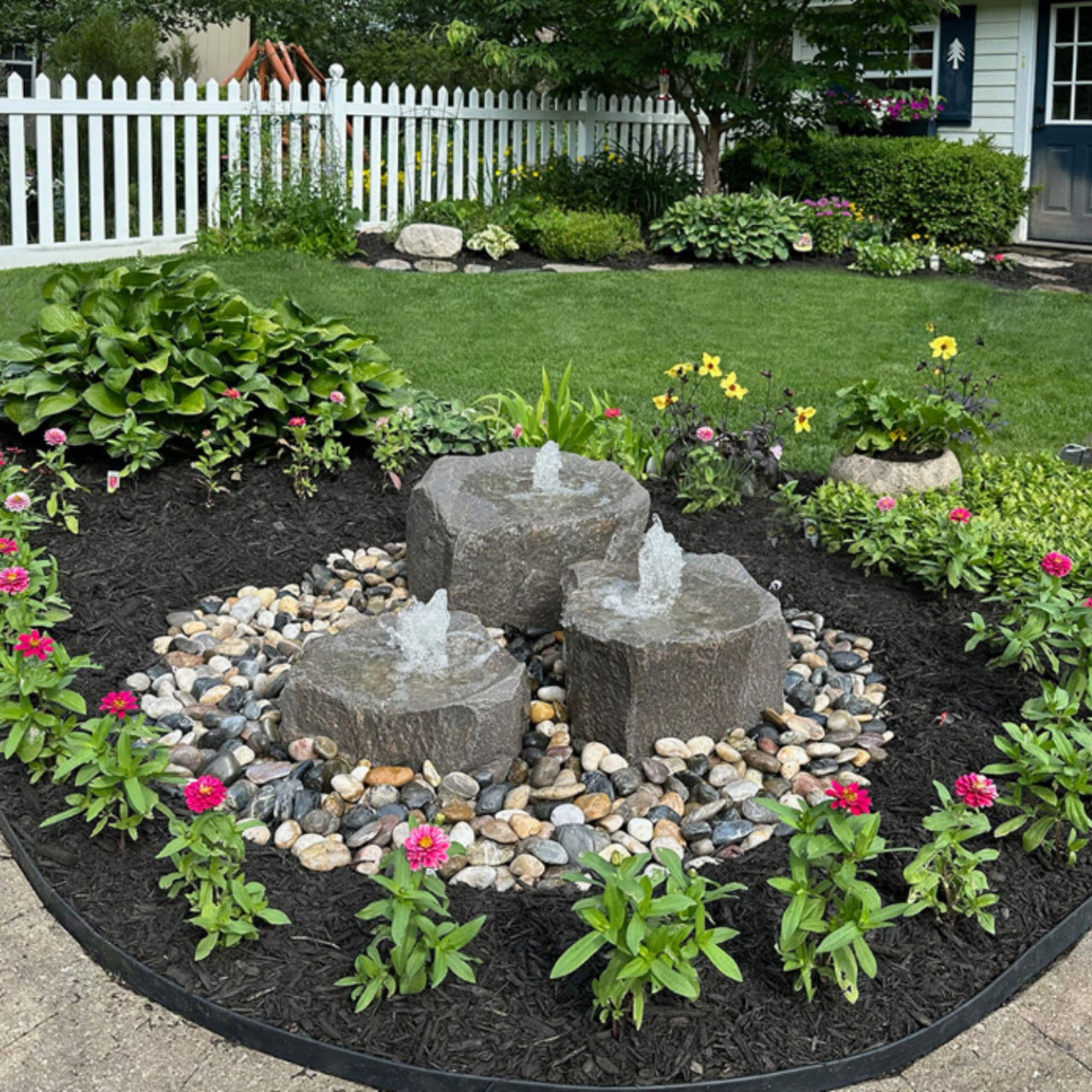 Triple Falls Garden Stone Fountain Surrounded By Flowers With White Fence Background
