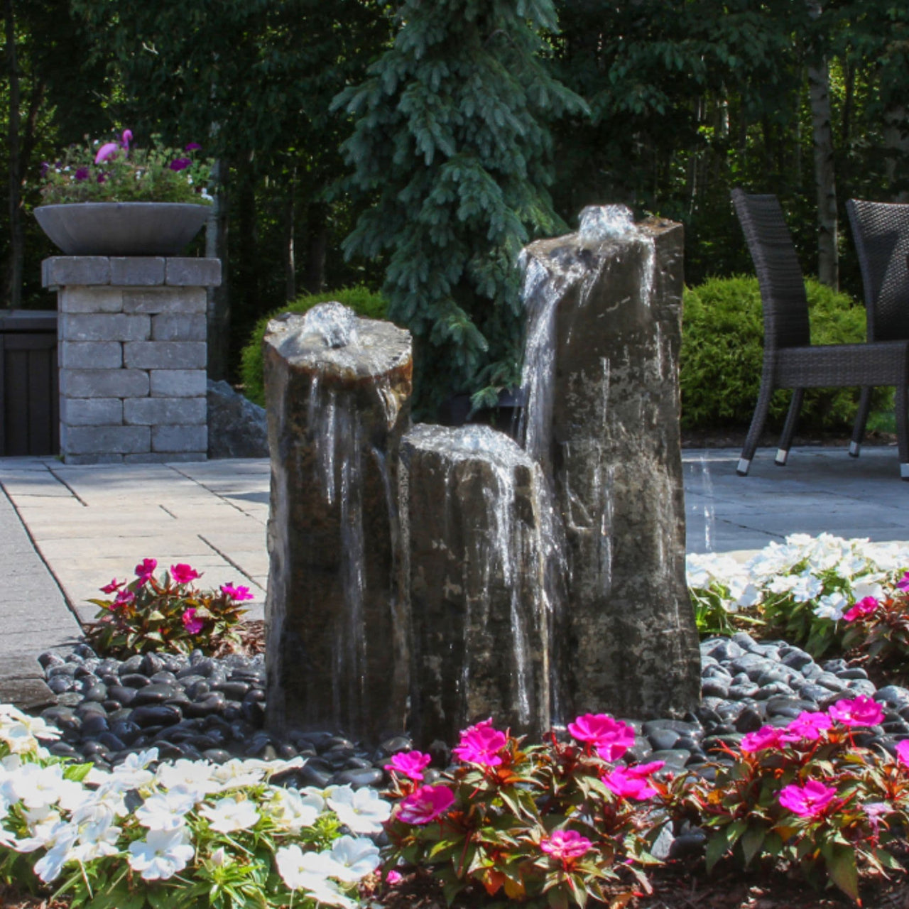 Triple Rustic Basalt Stone Outdoor Fountain Surrounded By Flowers With Trees Background