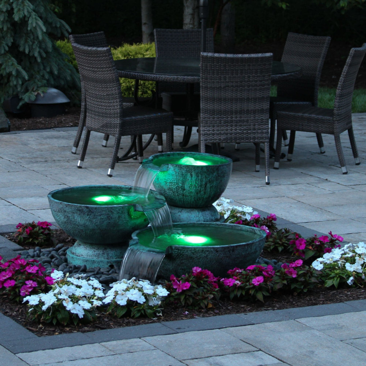 Triple Spillway Stone Bowl Fountain Surrounded By White And Pink Flowers With Table And Chairs Background