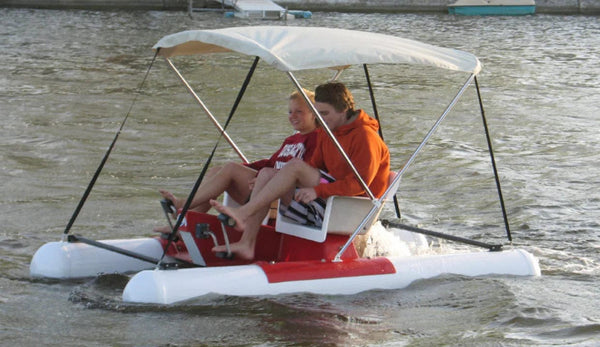 Red and white pontoon pedal boat with a white bimini sun canopy, carrying two riders across open water.